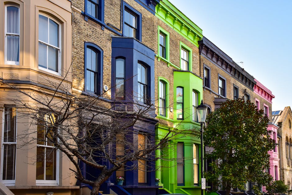 A row of three to four-storey terraced houses in Notting Hill, with ornate bay windows, decorative cornices, and small balconies. The buildings are painted in light pastel colours, primarily shades of