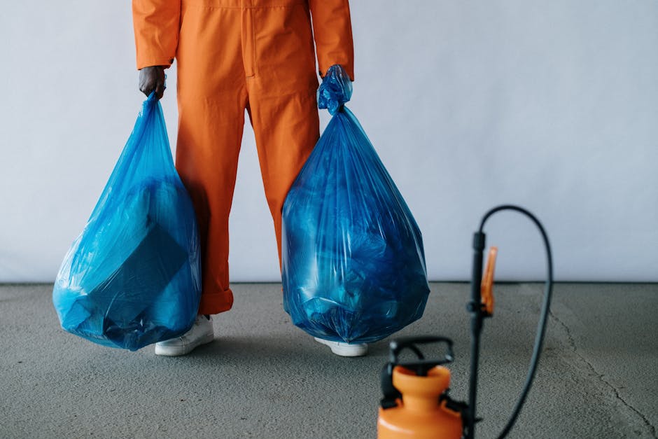 A person dressed in orange work overalls is standing indoors on a grey concrete floor, holding two large blue plastic garbage bags filled with waste, with one bag in each hand. Their face is not visib