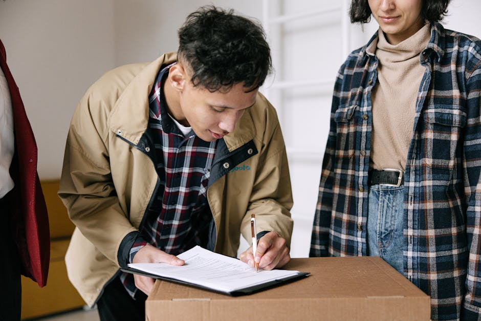 A person dressed in dark blue work overalls and pink slip-on shoes stands indoors against a light, textured wall, taking notes on a clipboard. Surrounding them are several cardboard boxes of varying s