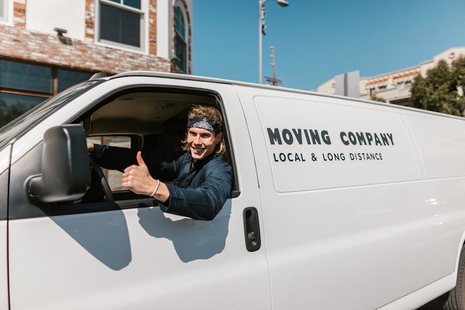 A man wearing a white t-shirt, dark jeans, and a cap walking beside a white cargo van parked on the street outside a residential property during daytime. The van is positioned parallel to the pavement