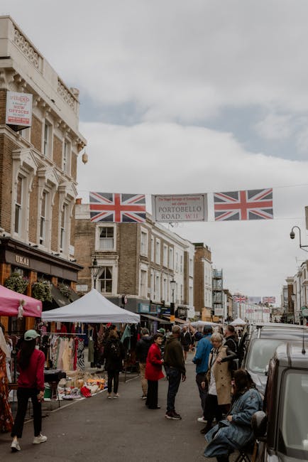 A busy street scene on Portobello Road in Notting Hill during a home relocation event, with several market stalls set up under colorful umbrellas displaying various goods on the sidewalk. pedestrians,